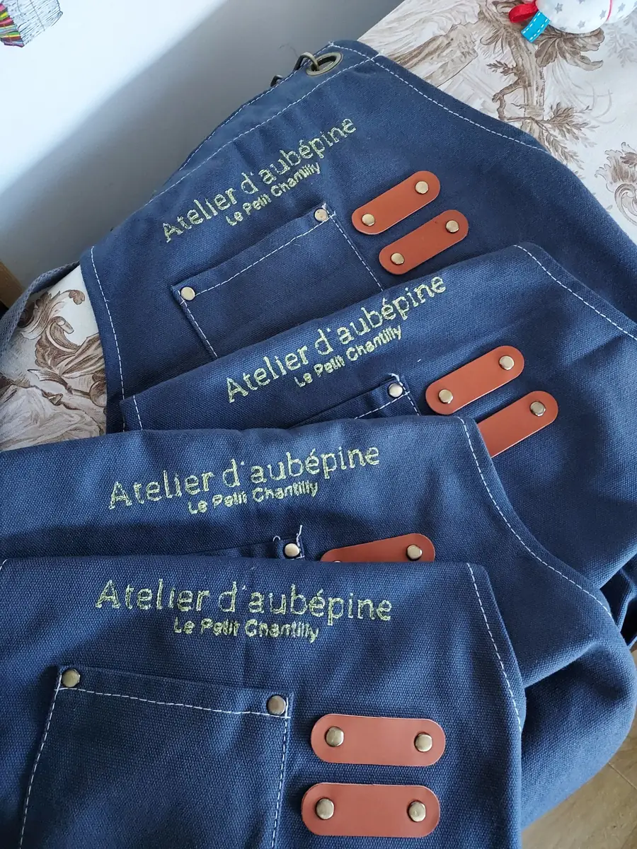 Quatre tabliers en toile bleue brodés au fil doré « Atelier d'aubépine – Le Petit Chantilly », avec une poche et des passants en cuir